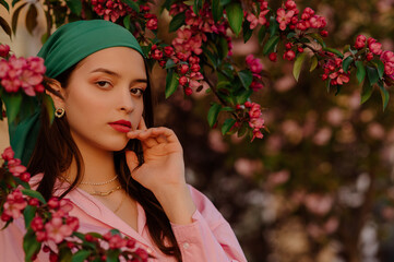 Fashionable woman wearing stylish green bandana, trendy earrings, posing near blooming tree....