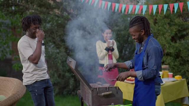 Laughing African American men talking cooking barbecue in summer garden with blurred teen girl drinking tea at background. Happy relaxed friends on picnic outdoors. Lifestyle concept