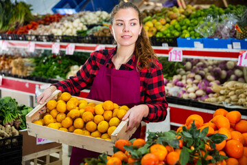 Young smiling woman salesman carrying box with fresh yellow plums at vegetable store