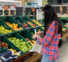Woman in a medical mask buying fruits in grocery supermarket. Protection from coronavirus epidemic, increased immunity with fresh fruit. Healthy food for fighting viruses.