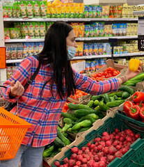 Woman in a medical mask choosing vegetables in a grocery supermarket. Protection from coronavirus epidemic, increased immunity with fresh fruit. Healthy food for fighting viruses.