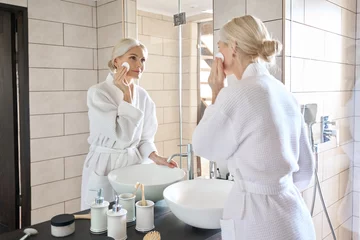 Foto op Canvas Senior mature older caucasian woman cleansing face with cotton pad removing makeup looking at mirror wearing bathrobe. Everyday routine, anti wrinkle prevention skin care products concept. © insta_photos