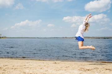 Happy woman in white shirt jumping on the sandy beach by the lake.