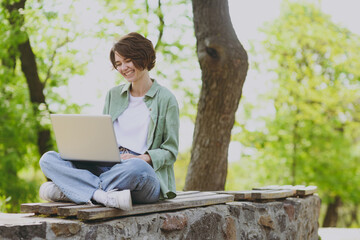 Full length young smiling happy fun student freelancer woman 20s in green jacket jeans sit on bench...