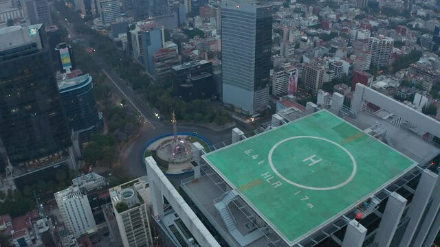 Drone camera flying over tall office building with green heliport on top. Aerial view of city near Angel of Independence monument. Mexico city, Mexico.