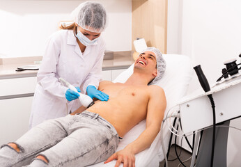 Laughing young man lying in cosmetology chair during hardware body procedure from professional...