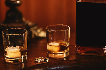 two glasses of cognac with ice, bottle with mockup and gold wedding rings on a wooden table. wedding day.