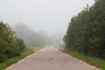 A deserted country road leading into the distance into the fog. Cracked asphalt on an old road. Trees and road signs on the roadside. Poor visibility in fog. Mystical mood.