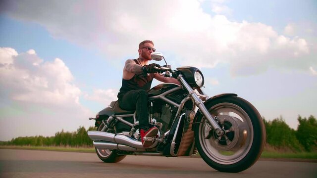 Stylish biker with tattoos rides a motorcycle on a country road at sunset