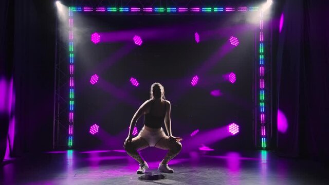Seductive sporty woman twerking ass in dark studio with colored lights. Silhouette of young woman in short white shorts and fishnet tights moving erotically with her body and hips. Slow motion.