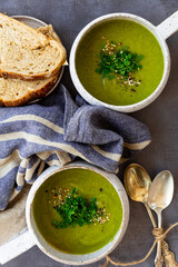 Green soup with toppings in white ceramic, napkins, spoon, bread slices on a gray surface