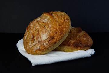 Two freshly baked loaves of homemade sourdough bread sit cooling on a clean white linen towel on an isolated black background.