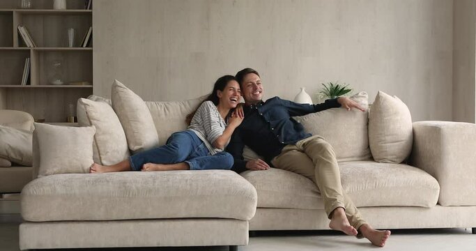 Young married couple talking laughing while resting on sofa. Spouses sit on comfy couch chatting look into distance, daydreaming having pleasant cheery conversation enjoy new home, relaxing on weekend