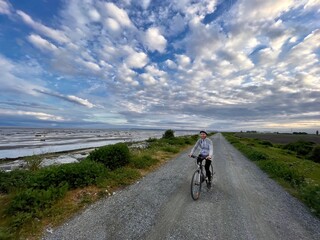 Person on a bike. Middle age woman cycling along the sea shore. Dyke trail in Delta. Vancouver. BC....