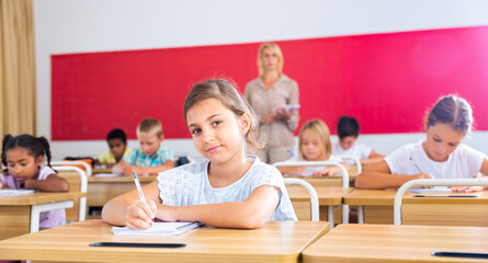 Fototapeta na wymiar Focused girl sitting at desk writing test in classroom full of pupils during lesson