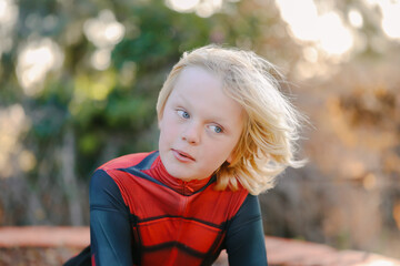 Little boy with long blonde hair sitting in golden sunlight wearing superhero costume