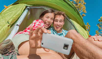 Camping selfie fun couple tourists holding phone taking self portrait photos in tent during summer vacation travel. Asian girl, Caucasian man laughing doing silly faces.