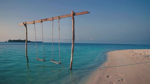 Cinemagraph seamless video loop of a wooden swing on a tropical remote dream honeymoon beach at the seaside on an empty natural adventure island in Indonesia with white sand by sunset. 4K UHD