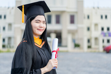 A young beautiful Asian woman university graduate in graduation gown and mortarboard holds a degree certificate stands in front of the university building after participating in college commencement