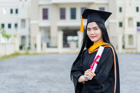 A young beautiful Asian woman university graduate in graduation gown and mortarboard holds a degree certificate stands in front of the university building after participating in college commencement