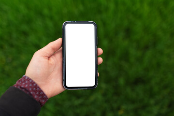 Close-up of male hand holding smartphone with white mockup on background of blurred green grass.