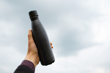 Close-up of male hand holding metal bottle on background of blurred clouds.