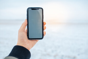 Close-up of male hand holding smartphone with mockup on background of blurred snowy field. Sunlight effect.