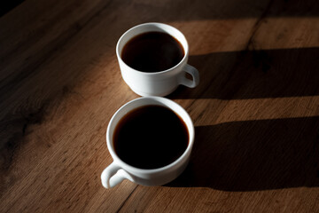 Close-up of two white ceramic cups of coffee with shadows on wooden table.