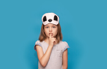 Cute little girl wearing sleep mask over blue isolated background ask to be quiet with finger on...