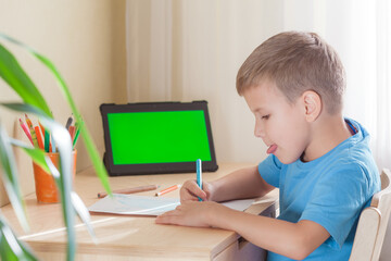 Smart school boy writing homework at desk in room, tablet with green screen stay on desk. Education and distance learning concept. - Powered by Adobe