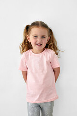 Adorable cheerful little girl with ponytails wearing pink t shirt standing on white background and looking at camera with smile