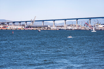 a section of the Coronado Bridge in San Diego showing shipyards under the bridge with mountains in the background and boats in the San Diego Bay in the foreground