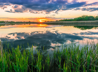Fototapeta na wymiar Scenic view at beautiful spring sunset with reflection on a shiny lake with green reeds, bushes, golden sun rays, calm water ,deep blue cloudy sky and glow on a background, spring evening landscape