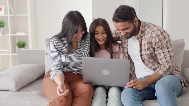 Parents with teenage daughter watching funny videos using laptop browsing online tv streaming enjoying spending time together on weekend at home in living room sitting on sofa. Happy indian family.