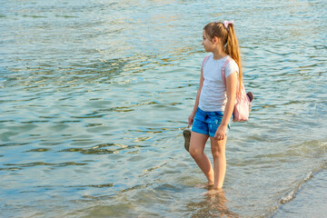 A cute teenage girl in shorts and a T-shirt barefoot walks along the coast in the golden ray of the...