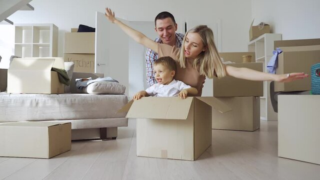 Overjoyed excited happy householders renters tenants parents celebrating the moving day with kid ride in carton box in new living room, having fun in their first real estate property. Slow motion.