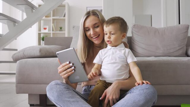 Young mom sitting at home on floor with kid of 3-4 years old, watching online activities classes for early education on digital tablet, or movies series for kids together.