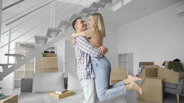 Happy young family couple embracing kissing in new home relocation. Joyful family in living room at moving day, boxes on background, first time home owners concept. Slow motion.