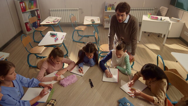 Students sitting at round table in classroom. Teacher explaining task of lesson