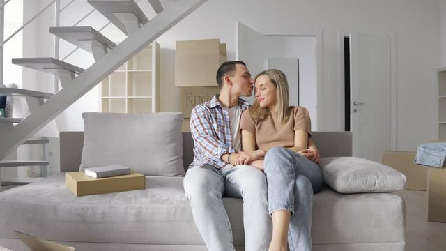 Happy young family couple enjoying time together embracing kissing in new home relocation sitting on sofa. Joyful family in living room at moving day, mortgage loan, first time buyers concept.