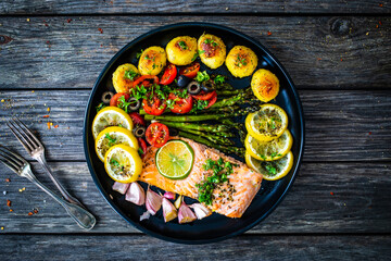 Roasted salmon steak with roast potatoes and vegetable salad served on black plate on wooden table
