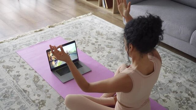 Young healthy African American mixed race girl wearing sportswear meditate watching online yoga class tutorial on pc at home, sit on mat doing fitness exercise feeling no stress balance harmony.