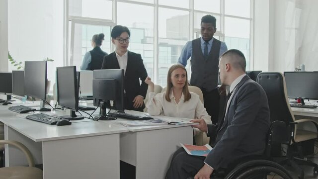 Tracking shot of disabled executive in wheelchair discussing work with male and female colleagues in office
