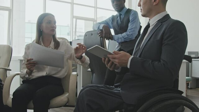 Tilt up of disabled executive in wheelchair showing something on tablet to his male and female colleagues in office while discussing work with them