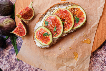 Figs. Sandwiches with figs, cheese, arugula, honey and nuts on wooden board over textured purple backdrop. Next to pieces of fresh figs with cinnamon. Homemade. Everyday autumn kitchen.