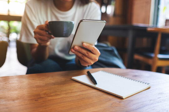 Closeup image of a woman holding and using mobile phone while drinking coffee with notebook on wooden table