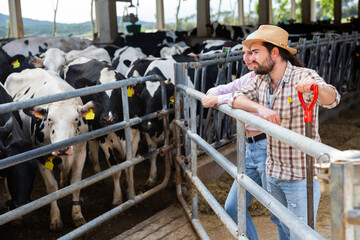 Confident young man and woman owners of dairy farm standing in stall on background with herd of cows..