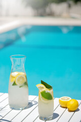 Homemade lemonade in glasses with lemon slice, on white table,in the edge of the pool with turquoise blue water background , light and shadows, vocation 2021 and summertime concept,space for text.