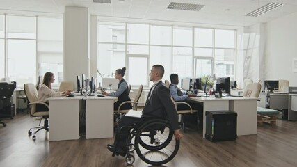 Wide shot of disabled businessman in wheelchair riding through open-plan office. Caucasian businesswomen and African-American businessman working on computers - Powered by Adobe