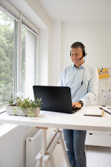 Man stands behind high table and speaks with headset and is working on laptop, notebook, pc